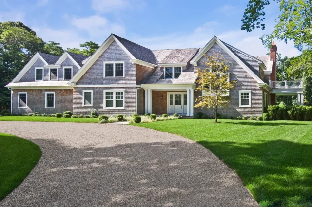Large modern suburban house with shingle siding, multiple gables, and a curved driveway surrounded by green lawn.