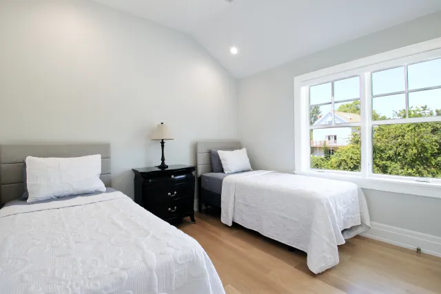 Bright bedroom with two twin beds, white bedding, black nightstand, lamp, and large window with greenery outside.