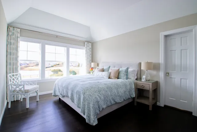 Bright modern bedroom with large window, light blue bedding, bedside tables, and white chair on dark wood floor