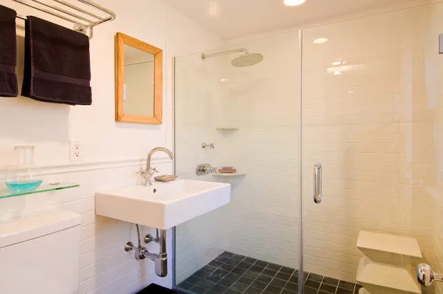 Modern bathroom with glass shower enclosure, white sink, wooden mirror, and black towels hanging on a rack.