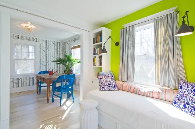 Bright living room with lime green wall, white daybed with patterned pillows, and adjacent dining area with blue chairs.