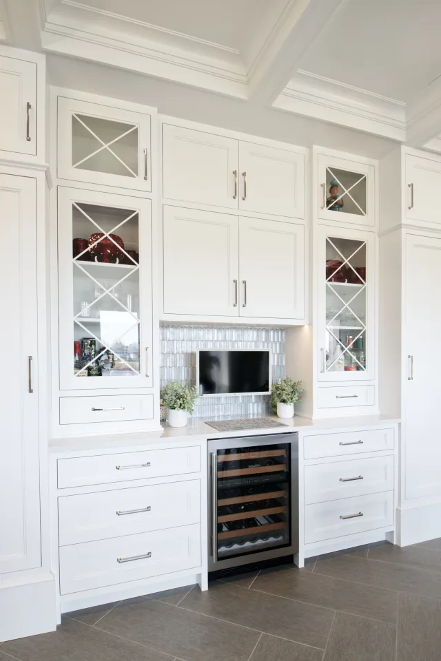 Modern white kitchen cabinetry with glass doors, built-in wine cooler, small TV, and decorative plants.