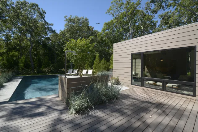 Modern backyard with wooden deck, pool, outdoor shower, and large windows overlooking a forested area