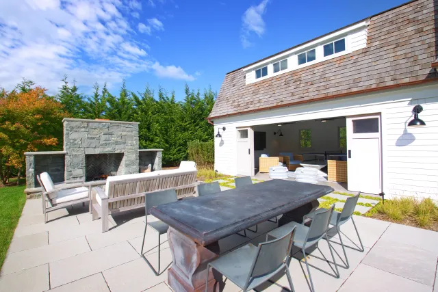 Modern outdoor patio with gray dining table, stone fireplace, white seating, and a bright blue sky.