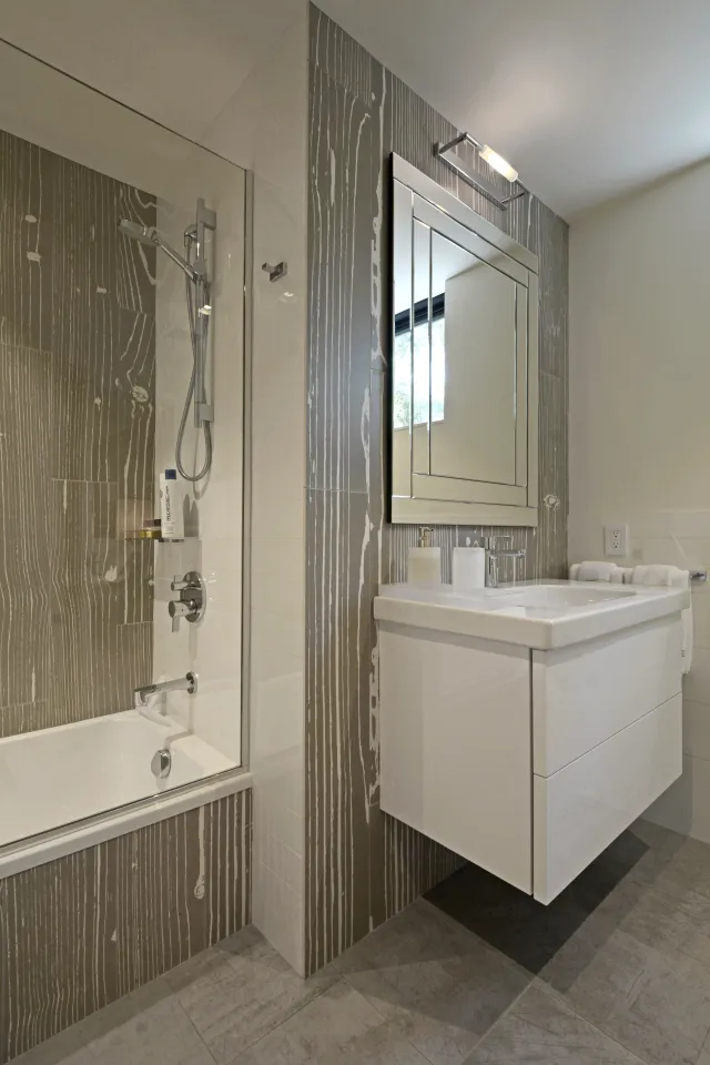 Modern bathroom with white floating vanity, large mirror, bathtub with glass shower door, and wood-patterned tile walls.