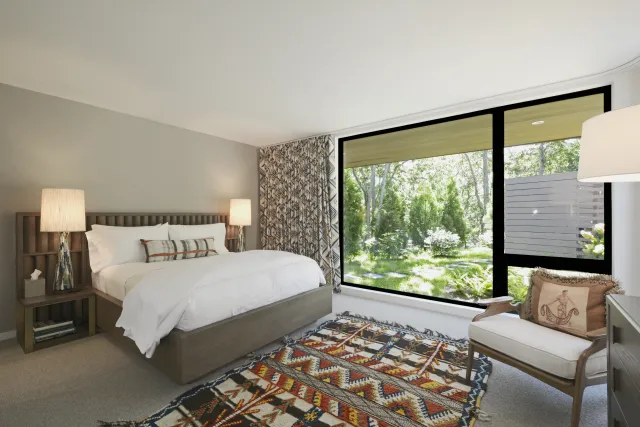 Modern bedroom with large window, wooden bed, patterned rug, and cozy armchair with pillow.