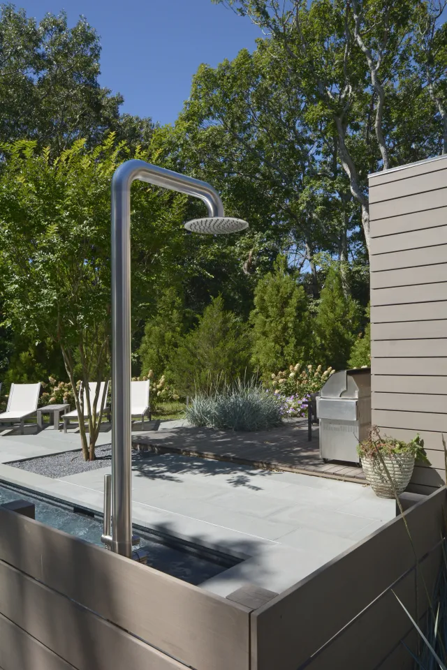 Outdoor shower next to a pool with lounge chairs, greenery, and a barbecue grill on a sunny day.