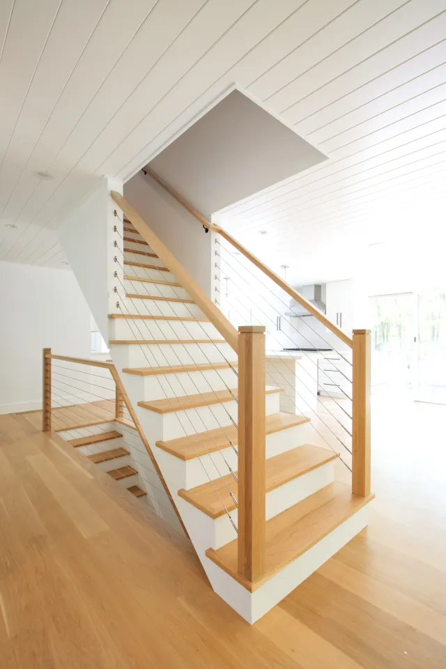 Modern wooden staircase with cable railing in bright open living space with light hardwood floors and white walls.