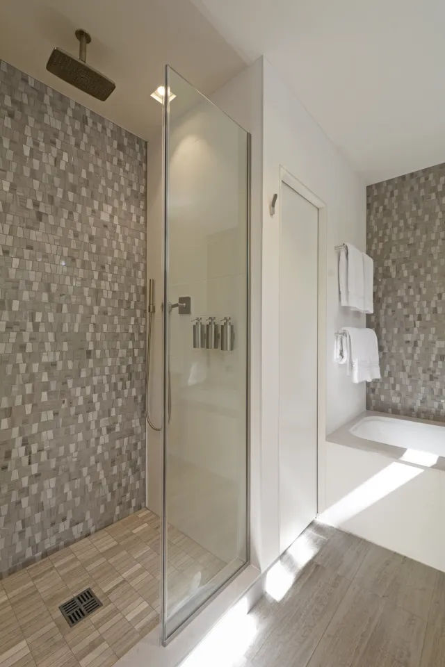 Modern bathroom with glass shower enclosure, gray tile walls, large showerhead, and white bathtub with towels.