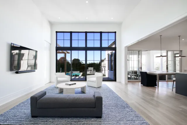 Modern living room with large black-framed windows, gray sofa, white chairs, and open dining area with pendant lights.
