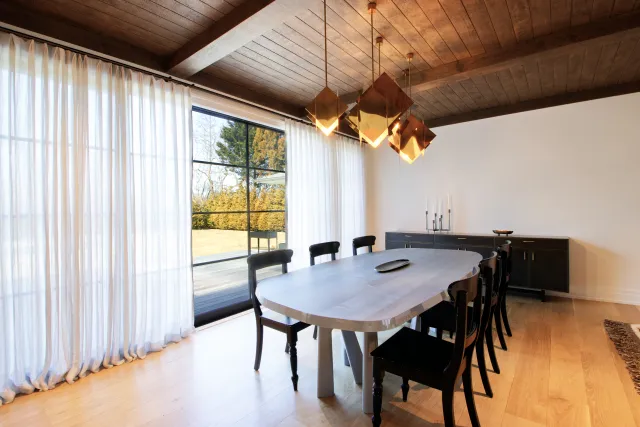 Modern dining room with wooden table, black chairs, gold pendant lights, hardwood floor, and large window with sheer curtains