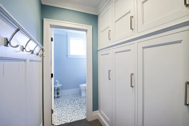Bright hallway with white cabinetry and hooks leading to a light blue bathroom with patterned tile floor.