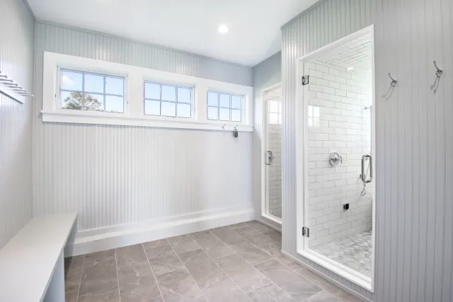 Bright white mudroom with gray tile floor, beadboard walls, bench seating, and glass door shower area.