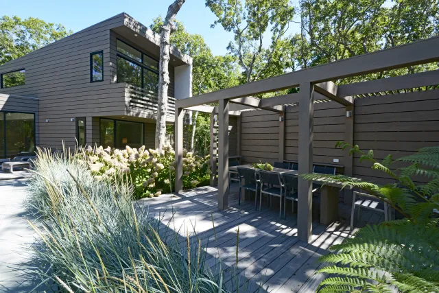 Modern house with outdoor patio featuring pergola, dining table, wooden deck, and surrounding greenery in daylight