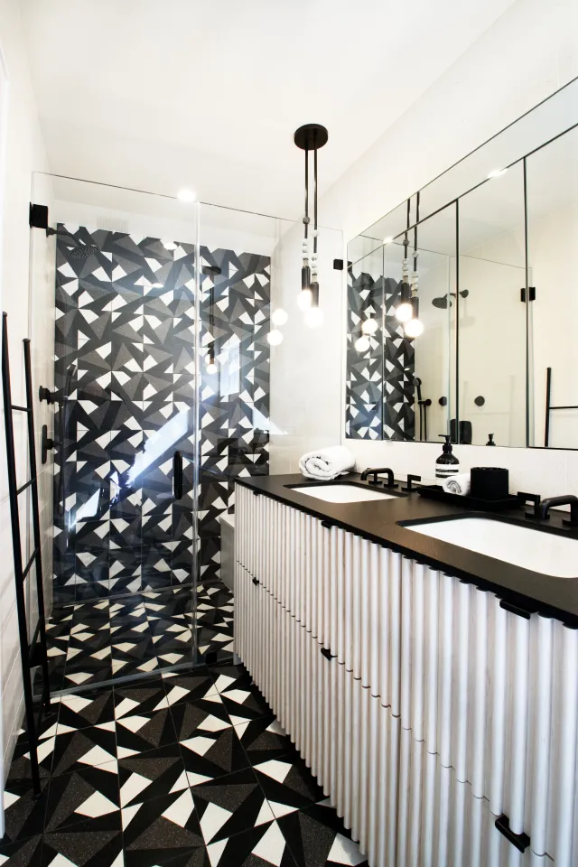 Modern bathroom with geometric black and white tiles, double sinks, frameless glass shower, and minimalist decor.