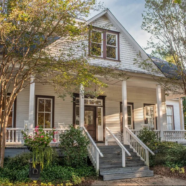 White wooden house with front porch, stairs, and greenery under a clear sky during daytime