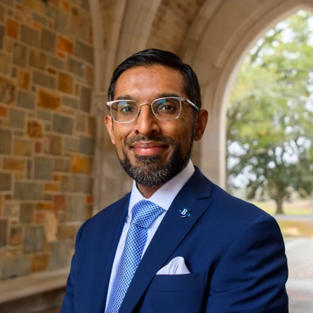 Man in blue suit and glasses standing under a stone archway with trees in the background outdoors.