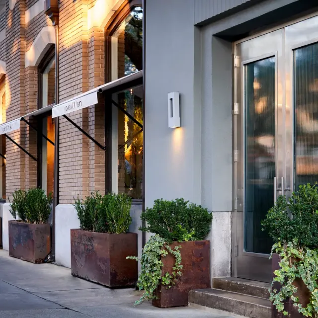 Modern storefront with large glass doors, brick walls, and rust-colored planters with green shrubs.