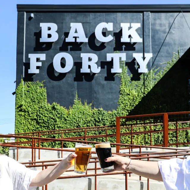 Two people clinking glasses of beer in front of the Back Forty sign amidst greenery.