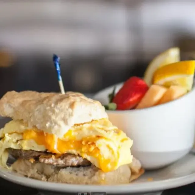 Breakfast sandwich with sausage, scrambled eggs, melted cheese on a biscuit, served with mixed fresh fruit.