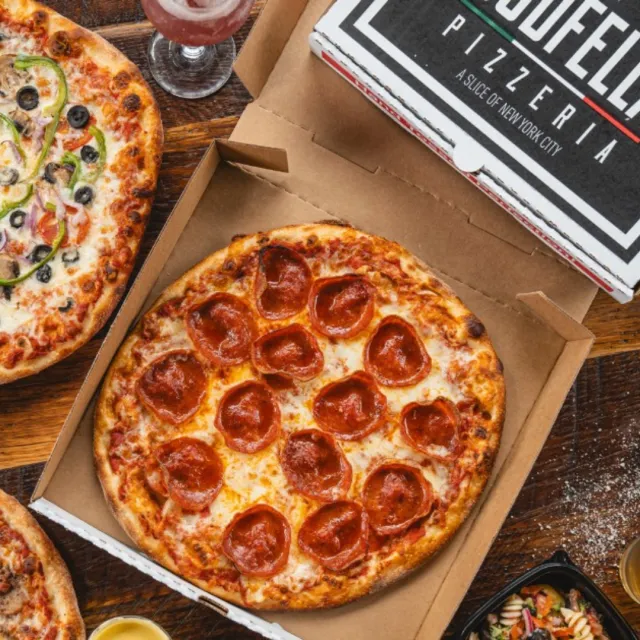 Pepperoni and vegetable pizzas in boxes on wooden table with salad and drinks, ready to serve.