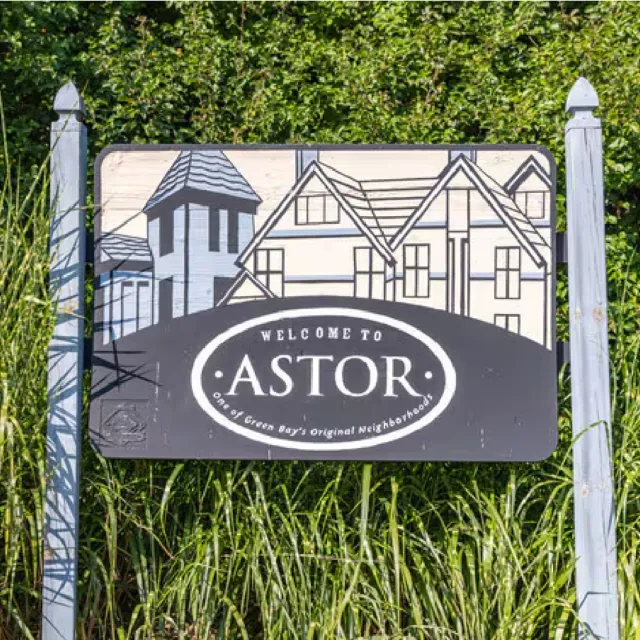 Welcome to Astor neighborhood sign with house illustrations surrounded by greenery and posts.
