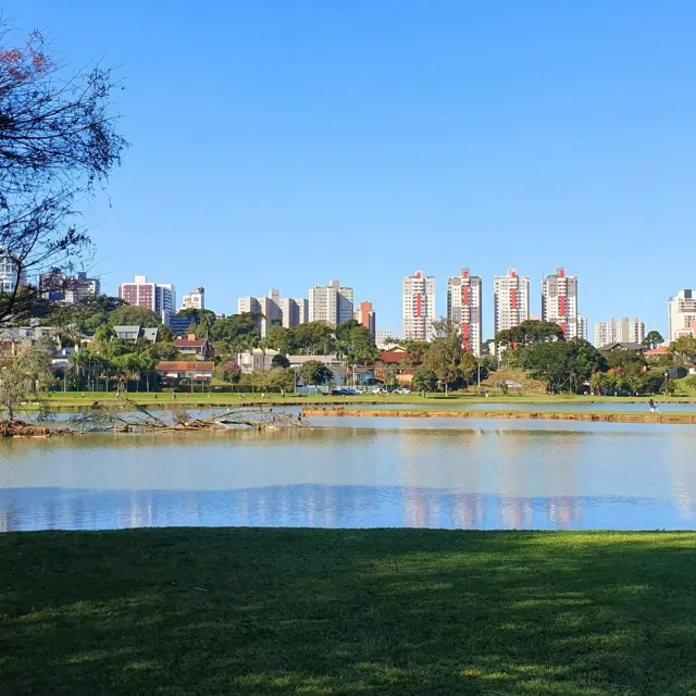 City skyline behind a calm lake with green grass and trees under a clear blue sky.