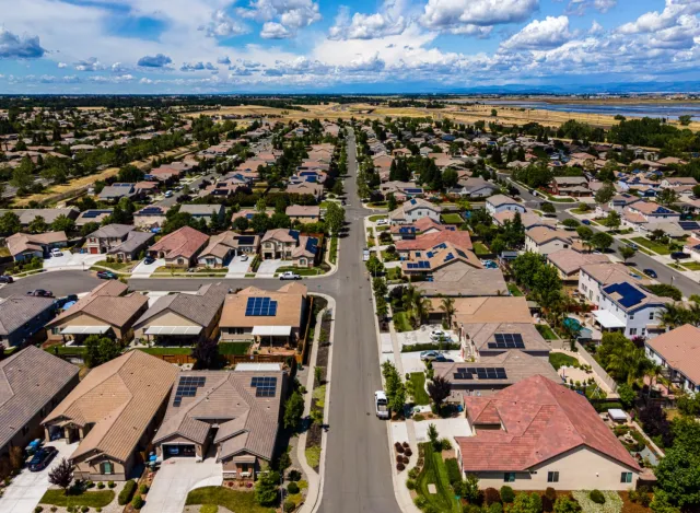 Aerial view of a suburban neighborhood with solar panels on many house roofs under a blue sky with scattered clouds.