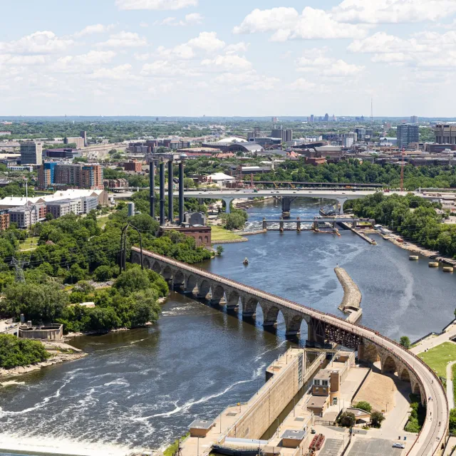 Aerial view of a city river with bridges, dam, greenery, and urban buildings under a blue sky.