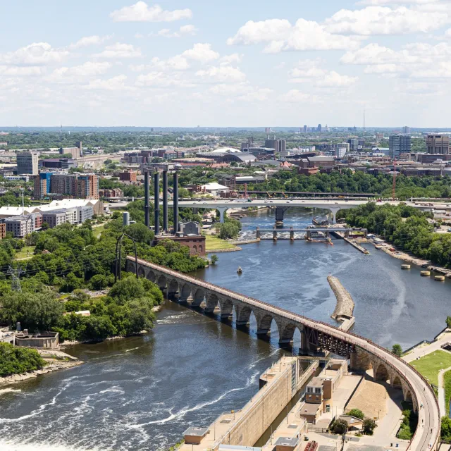 Aerial view of a river with bridges, a dam, green trees, and a cityscape under a partly cloudy sky.