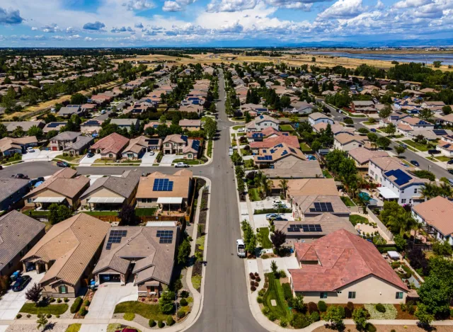 Aerial view of suburban neighborhood with solar panels on rooftops under a blue sky with white clouds.