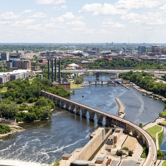 Aerial view of a river with a historic stone arch bridge, dam, and urban cityscape under a blue sky.