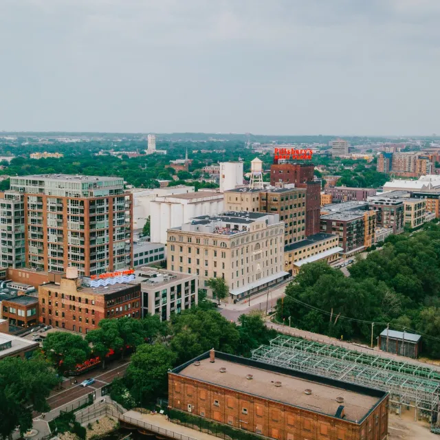 Aerial view of an urban area with mid-rise buildings, dense greenery, and a cityscape under a cloudy sky.