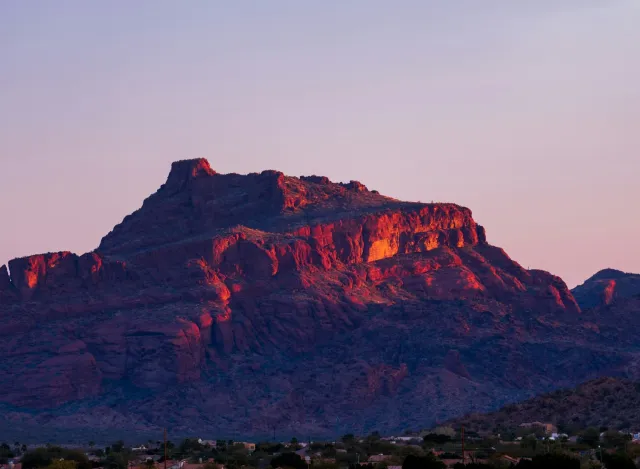 Sunset casts warm red and orange hues on a rugged mountain range above a desert town landscape.