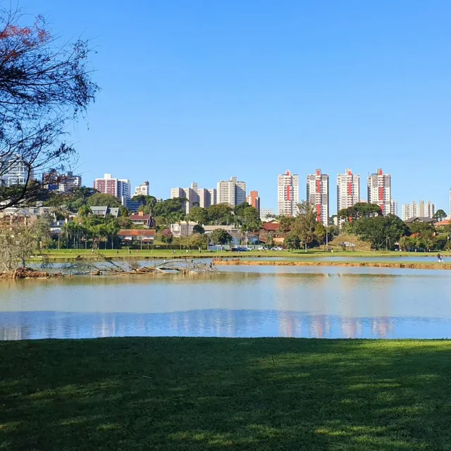 City skyline with tall buildings across a calm lake under a clear blue sky and grassy park foreground
