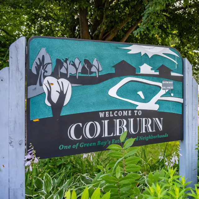 Welcome sign for Colburn Neighborhood in Green Bay featuring trees, houses, and a pond illustration.