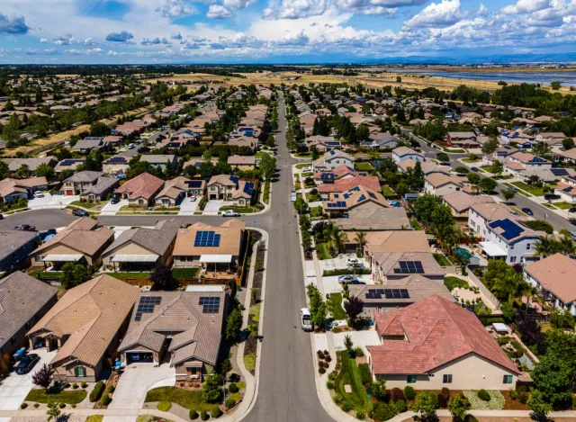 Aerial view of a suburban neighborhood with solar panels on roofs under a blue sky with scattered clouds.