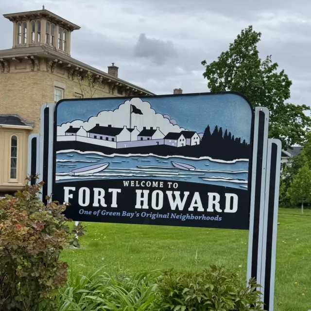Welcome sign for Fort Howard in Green Bay featuring historic buildings and boats by the water under a cloudy sky.
