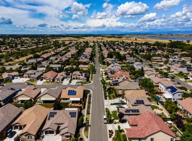 Aerial view of suburban neighborhood with solar panels on rooftops under a partly cloudy blue sky.