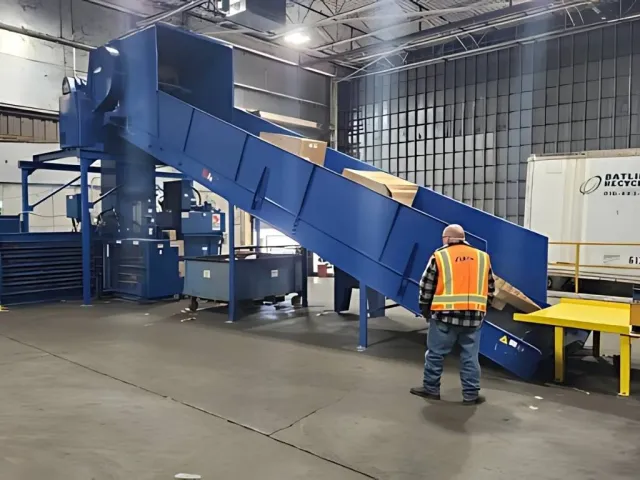 Worker in safety vest operating a blue conveyor belt carrying boxes inside a warehouse facility.