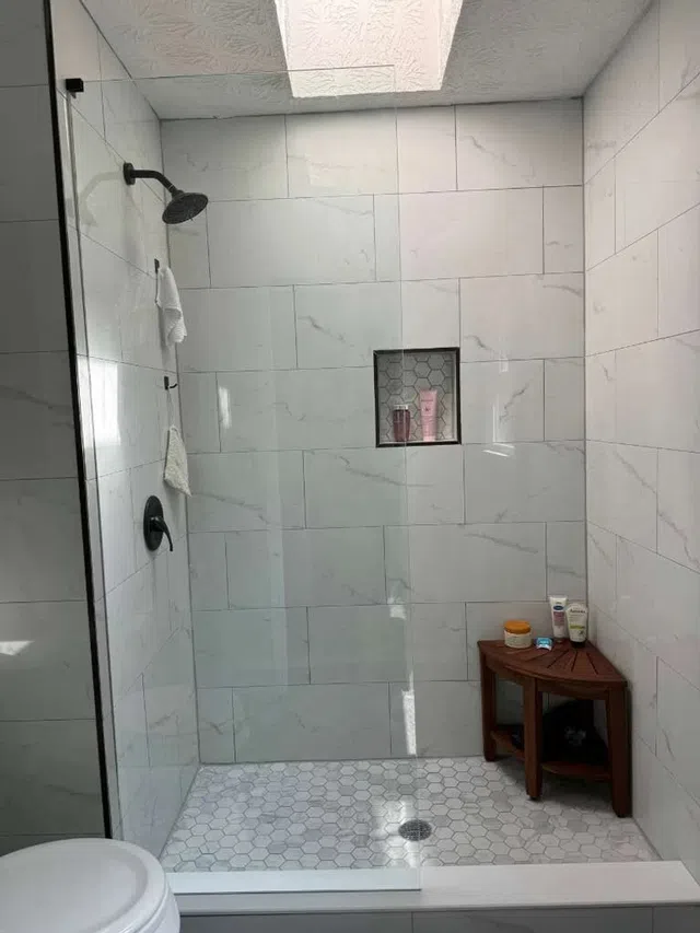 Modern walk-in shower with large white marble tiles, hexagonal floor tiles, skylight, and wooden corner bench.