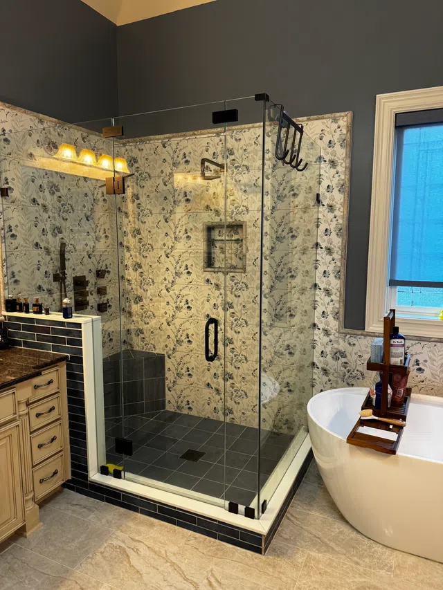 Modern bathroom with glass shower, floral tile walls, freestanding tub with wooden tray, and dark blue accents.