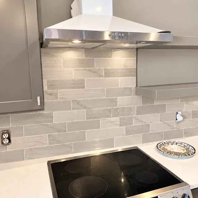 Modern kitchen stovetop with digital controls, stainless steel range hood, gray tile backsplash, and white countertop.
