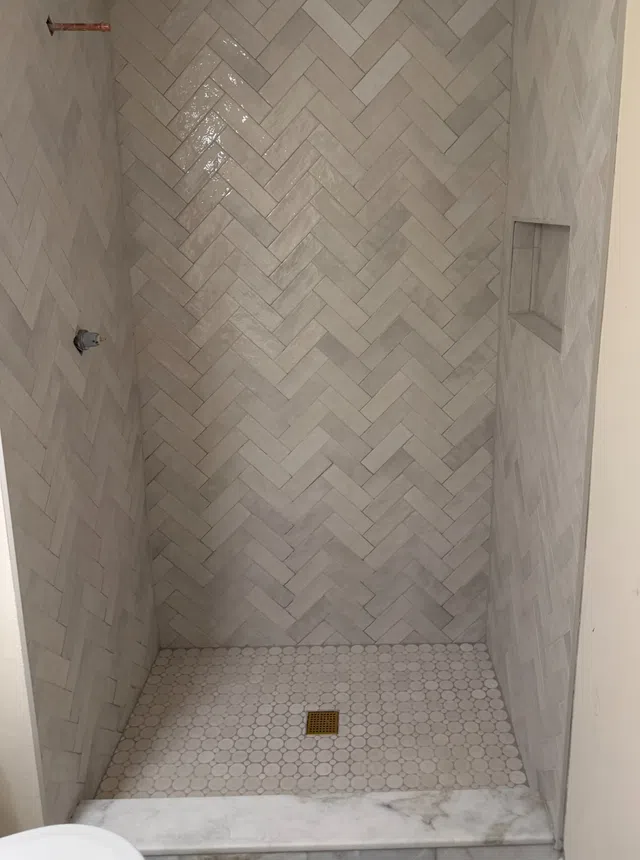 Shower with light grey herringbone wall tiles and round mosaic floor tiles with brass drain cover