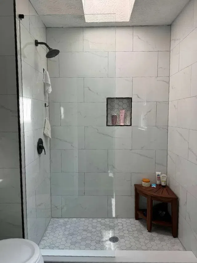 Modern walk-in shower with large white marble tiles, hexagonal floor tiles, skylight, and wooden corner bench.