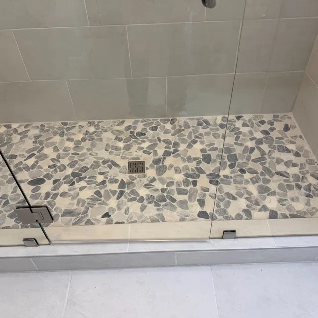 Modern shower area with pebble stone floor tiles and glass door enclosure with metal hinges and drain.
