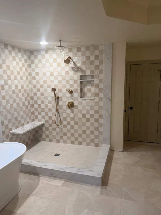Modern walk-in shower with beige and white checkerboard tiles, bronze fixtures, built-in shelf, and marble bench.