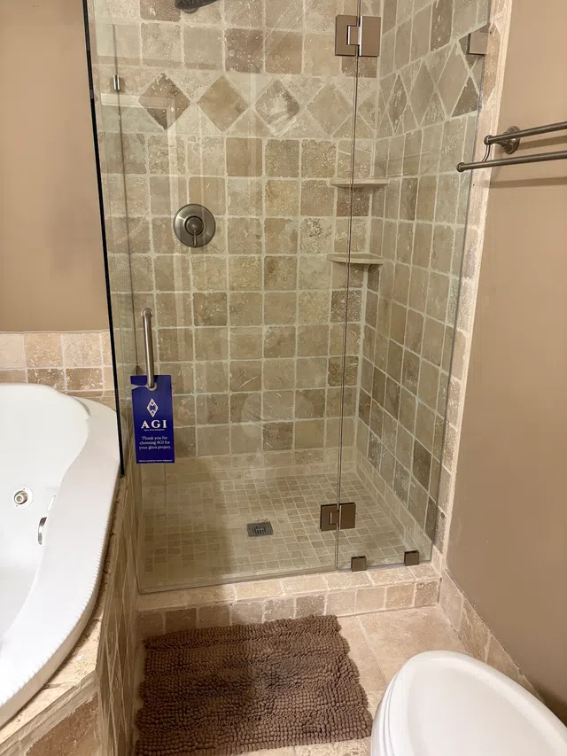 Glass shower enclosure with beige tile walls, built-in shelves, and a brown bath mat next to a white bathtub.