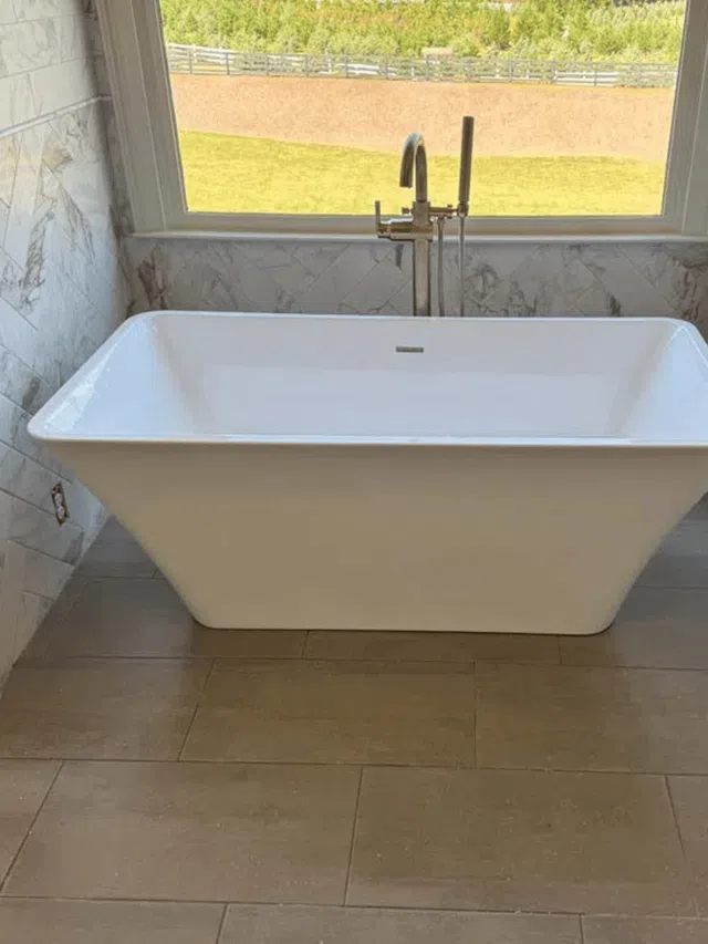 Modern white freestanding bathtub with floor-mounted faucet against marble tile wall and large window.