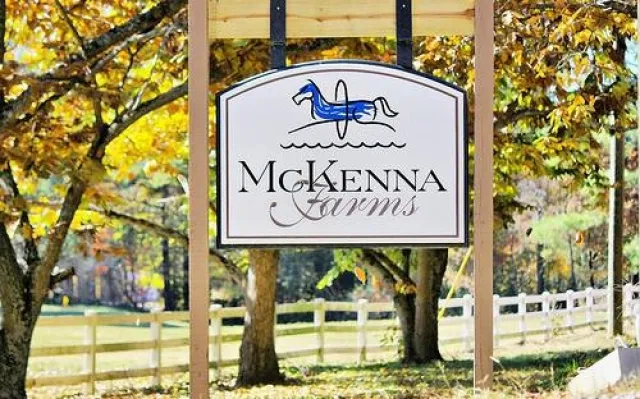 McKenna Farms wooden sign with blue horse logo hanging outdoors by trees and white fence in autumn setting